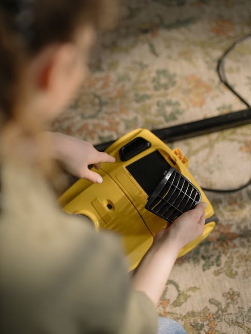A person, viewed from above and slightly obscured, is operating a yellow portable air mover with black vents, positioned on a patterned, multicolored carpet in a room with a wooden floor. The room appears well-lit, and the carpet shows no visible dirt or stains, suggesting recent deep cleaning or drying. The individual’s hands are on the air mover, which is part of a sanitisation or drying process, with a black power cable extending into the background. This scene reflects professional cleaning activity, likely undertaken by Carpet Cleaning Kingston, as part of surface cleaning or hygienic maintenance in a domestic setting.