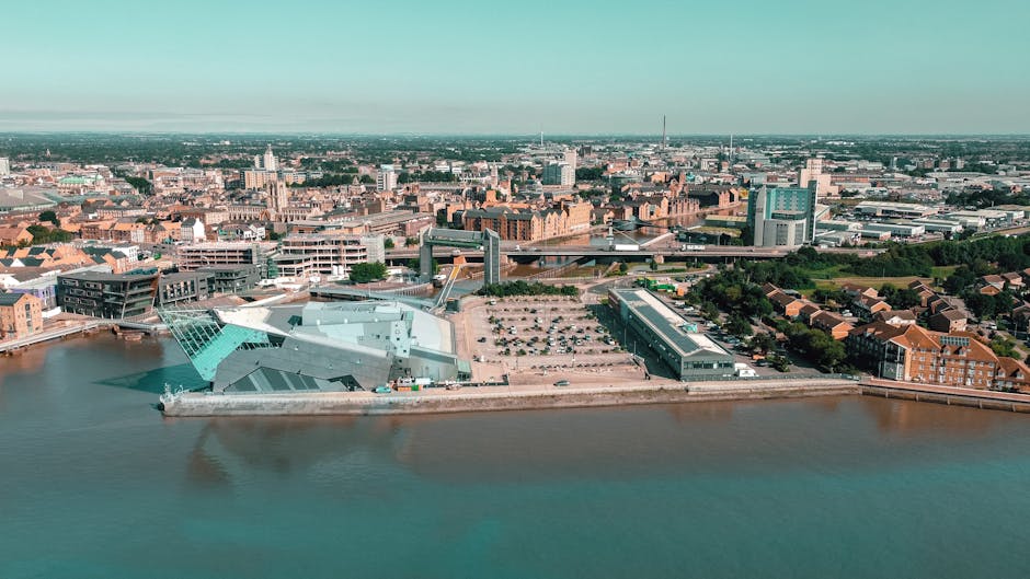 Aerial view of Kingston Riverside near Kingston Bridge, showcasing modern and historic buildings along the riverbank, including the contemporary glass-structured Kingston Museum and the adjacent complex with a large parking lot. The river expands across the foreground, reflecting the sky, with the surrounding urban area featuring a mix of brick and glass facades, and green spaces interspersed among the buildings. The scene is illuminated by natural daylight under a clear sky, highlighting the clean, well-maintained surfaces of the river, pathways, and buildings, reflecting the importance of hygiene and regular surface cleaning. Carpet Cleaning Kingston would recommend ensuring such environments are regularly deep cleaned and sanitised to maintain hygiene standards in both domestic and commercial spaces.