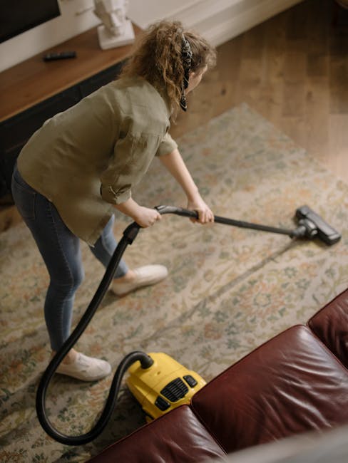 A woman using a vacuum cleaner on a patterned area rug in a living room, with a brown leather sofa in the foreground and a wooden sideboard with decorative items along the wall. The room has warm lighting and a polished wooden floor visible near the rug. The image illustrates professional carpet cleaning processes, highlighting surface cleaning and deep cleaning techniques associated with residential sanitisation, as provided by Carpet Cleaning Kingston near Kingston Bridge. The woman is dressed casually, and no other cleaning tools or equipment are visible besides the vacuum cleaner, which appears to be a commercial model suitable for thorough cleaning of domestic carpets.