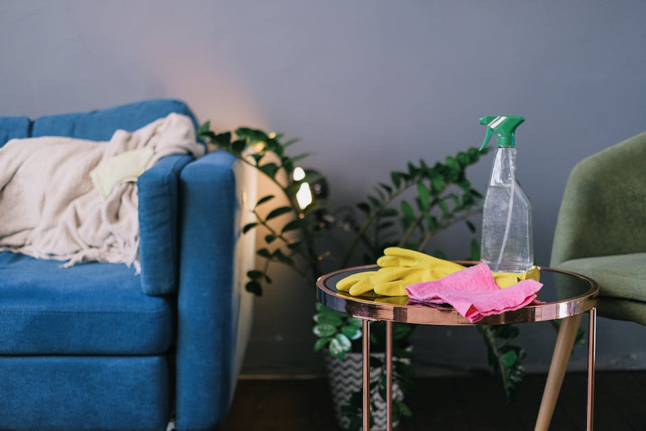 A close-up view of a living room corner in Kingston flats showing a worn blue fabric sofa with a cream blanket draped over its armrest. Next to the sofa, a small round side table with a metallic, rose gold finish holds yellow rubber gloves, a pink cleaning cloth, and a clear spray bottle filled with cleaning solution. Behind the table, a potted green plant adds a touch of freshness, and soft ambient lighting illuminates the space. The room has a tidy appearance with visible signs of recent cleaning activity, emphasizing the importance of regular surface cleaning and sanitisation as part of professional domestic cleaning services offered by Carpet Cleaning Kingston, especially when addressing common problems related to sofa cleaning in residential flats.