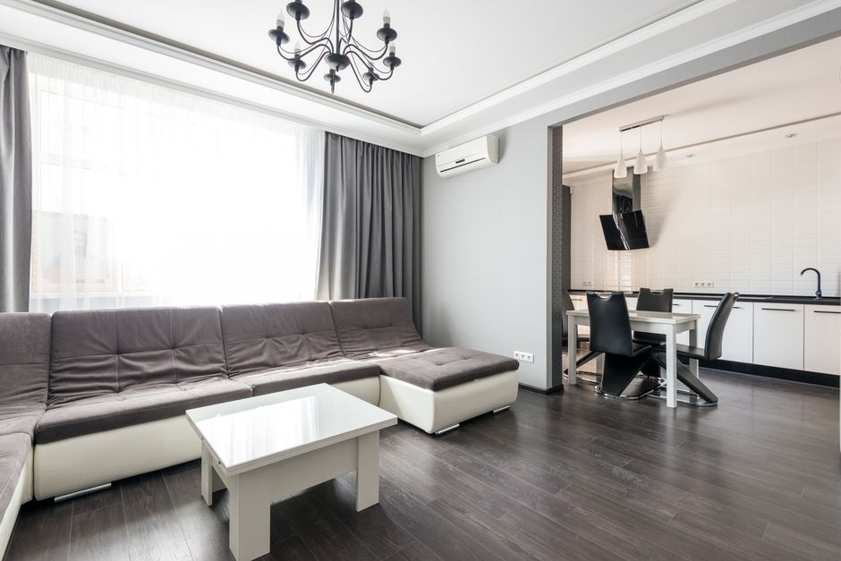 A modern living room in a Kingston flat featuring a large L-shaped sofa with a combination of dark grey fabric cushions and white base, positioned on dark wooden flooring. A white coffee table is placed in front of the sofa, and large windows with sheer white curtains and grey drapes allow natural light to brighten the space. An air conditioning unit is mounted on the wall above the sofa. Adjacent to the living area, an open-plan dining space includes a white table surrounded by four black chairs, with a kitchen featuring white cabinets, a black countertop, and a wall-mounted black range hood visible in the background. The room appears clean, well-maintained, and well-lit, highlighting the importance of surface cleaning and hygiene in domestic environments, as recent deep cleaning by Carpet Cleaning Kingston can help address access challenges in sofa cleaning within Kingston flats.