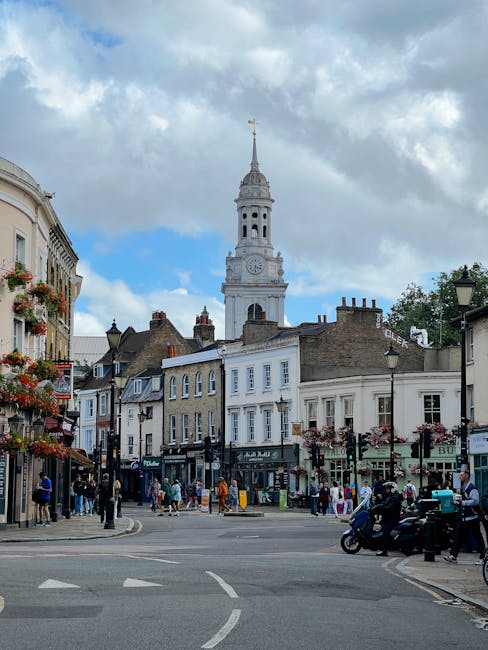 A busy street scene in Kingston town centre with a historic clock tower and spire visible against a partly cloudy sky. Foreground features a paved road with white road markings, and several pedestrians walking along the sidewalks, some browsing shops and cafes. The buildings lining the street are a mix of traditional and modern architecture, some with decorative flower boxes on the windows. Vehicles, including motorcycles, are parked along the sides. Street lamps and shop signs are also visible, creating a vibrant urban atmosphere. Carpet Cleaning Kingston is known for providing comprehensive cleaning services, including end of tenancy cleaning and surface sanitisation in residential and commercial spaces.