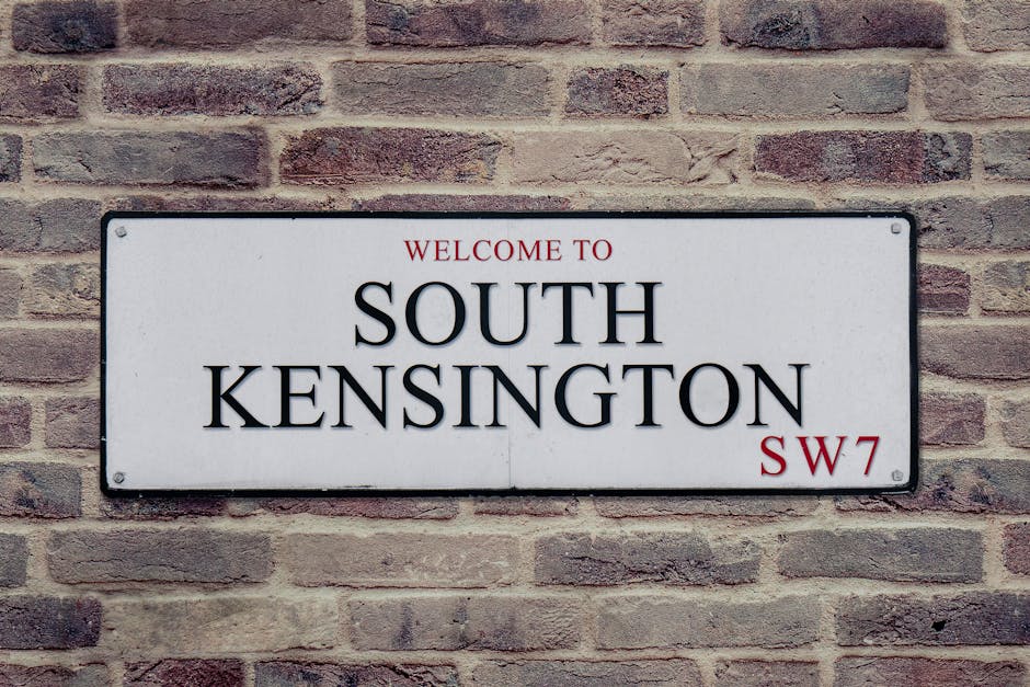 A rectangular white sign with black and red lettering mounted on a brick wall wall. The sign displays the words 'Welcome to South Kensington' with 'South Kensington' in large, bold black font, and 'Welcome to' and 'SW7' in smaller red font. The brick wall behind the sign features a traditional running bond pattern with reddish-brown bricks and light mortar joints, illuminated evenly by natural or ambient light, creating a clean and orderly appearance characteristic of a well-maintained urban environment. The sign and brick wall are free of dirt or damage, reflecting a focus on cleanliness and surface maintenance typical of professional cleaning standards by Carpet Cleaning Kingston.