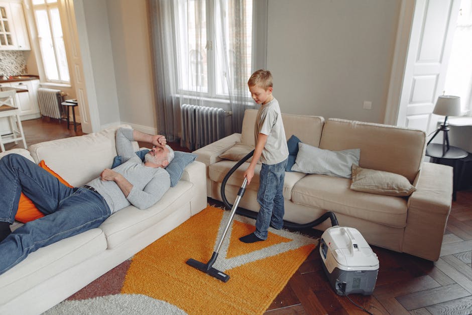 A young boy using a vacuum cleaner to perform deep surface cleaning on a light beige fabric sofa in a living room, while an older man with gray hair and beard reclines on the sofa, resting with his eyes closed. The room features wooden flooring with a multicolored area rug, large windows with curtains allowing natural light, and a side table with a lamp. The scene depicts domestic cleaning and sanitisation, showcasing professional cleaning processes that improve hygiene and maintain a clean, inviting environment, as promoted by Carpet Cleaning Kingston on their Kingston home service page.