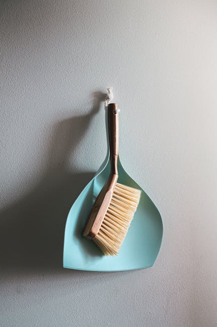 A dustpan and brush are hanging on a white wall in what appears to be a domestic setting. The dustpan is light blue with a smooth surface, and the brush has a wooden handle with beige bristles. The cleaning tools are positioned together, with the brush resting inside the dustpan, which is mounted on a wall hook. The wall surface is clean and unblemished, and the area is illuminated with soft, natural light, reflecting a tidy and organized environment suitable for surface cleaning, deep cleaning, or sanitisation in a home or commercial space. This setup suggests preparation for partial cleaning activities as part of regular maintenance, consistent with the guidance for landlords on cleaning compliance, as outlined by Kingston Council.
