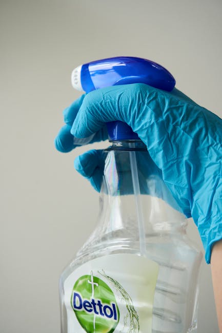 A close-up of a hand wearing a bright blue nitrile glove holding a clear plastic spray bottle filled with a light-colored cleaning solution. The spray nozzle is blue and white, and the bottle features a green 'Dettol' logo, indicating a disinfectant or sanitising product. The background is neutral and slightly blurred, emphasizing the cleaning product and gloved hand, which illustrates the process of surface sanitisation or deep cleaning, relevant to residential or commercial hygiene maintenance. The setting suggests professional cleaning practices, aligning with surface cleaning and sanitisation standards discussed in the Kingston Council guidance for landlords, as provided by Carpet Cleaning Kingston.