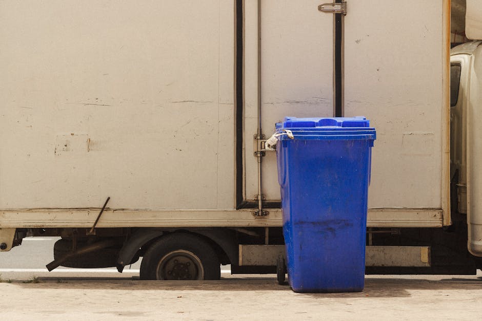 A blue recycling bin standing on a paved surface next to a white delivery truck with a closed metal door. The truck appears aged, with visible dirt and signs of wear on its surface. The bin is positioned close to the truck's side, with a handle on its lid. The scene is lit with natural daylight, highlighting the clean and orderly appearance of the bin in contrast to the slightly weathered condition of the truck. The environment suggests an outdoor setting, possibly a loading or unloading area, with the focus on the surface cleaning and waste disposal considerations. Carpet Cleaning Kingston emphasizes proper waste management in accordance with Kingston Council rules for disposing carpet waste after cleaning, within the context of domestic and commercial cleaning services.