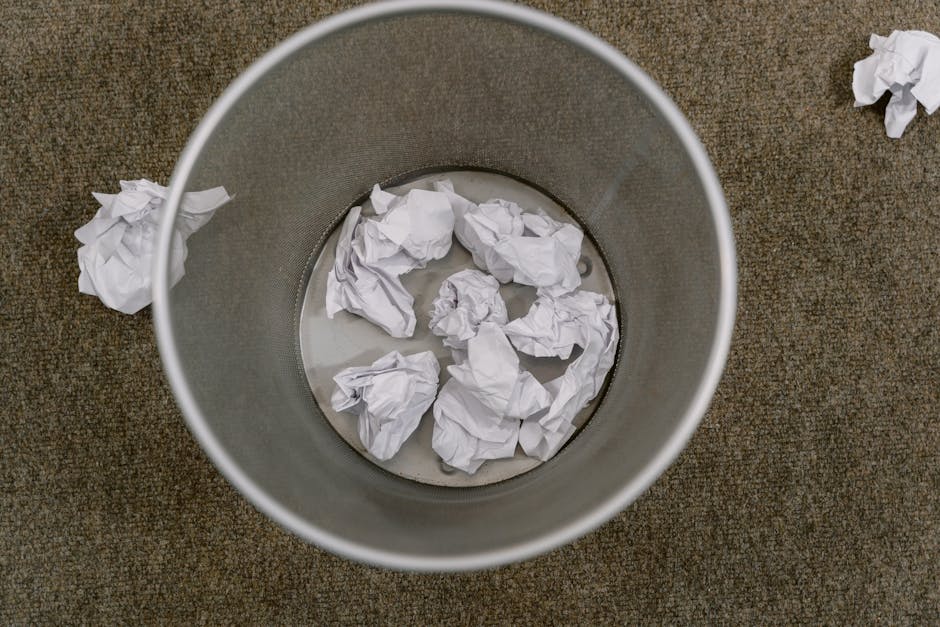A blue recycling bin standing on a paved surface next to a white delivery truck with a closed metal door. The truck appears aged, with visible dirt and signs of wear on its surface. The bin is positioned close to the truck's side, with a handle on its lid. The scene is lit with natural daylight, highlighting the clean and orderly appearance of the bin in contrast to the slightly weathered condition of the truck. The environment suggests an outdoor setting, possibly a loading or unloading area, with the focus on the surface cleaning and waste disposal considerations. Carpet Cleaning Kingston emphasizes proper waste management in accordance with Kingston Council rules for disposing carpet waste after cleaning, within the context of domestic and commercial cleaning services.