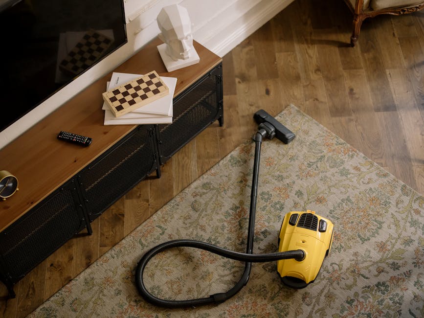A yellow vacuum cleaner with a black hose and silver wand attachment is positioned on a patterned area rug in a room with wooden flooring. To the left, a low black metal and wood media console holds a remote control, a white bust sculpture, and a stack of papers with a chessboard on top. The room features white crown molding along the wall, which is painted in a neutral tone, and the lighting highlights the clean, polished surface of the wooden floors and the well-maintained condition of the rug. The scene depicts a domestic setting focused on surface cleaning and maintenance, with emphasis on thorough cleaning tools used by Carpet Cleaning Kingston for deep cleaning and sanitisation services.