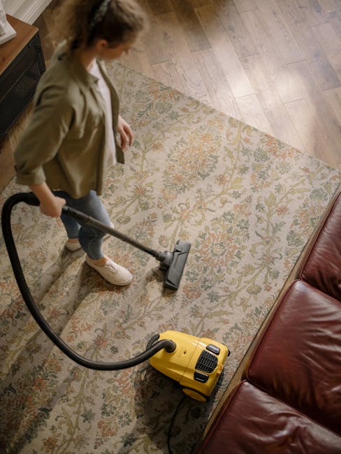 A woman is engaged in surface cleaning with a vacuum cleaner in a residential living room. She is standing on a patterned area rug with floral motifs in shades of beige, green, and orange, laid over a wooden floor. The room is naturally lit, highlighting the clean and well-maintained condition of the space. Nearby, there is a red leather sofa, and the woman is dressed casually, wearing a beige jacket, jeans, and sneakers. The vacuum cleaner is a bright yellow model, visibly in use for deep cleaning of the carpet and surrounding flooring. The scene reflects professional domestic cleaning or routine maintenance aimed at maintaining hygiene and cleanliness, aligned with the services promoted by Carpet Cleaning Kingston in Kingston.
