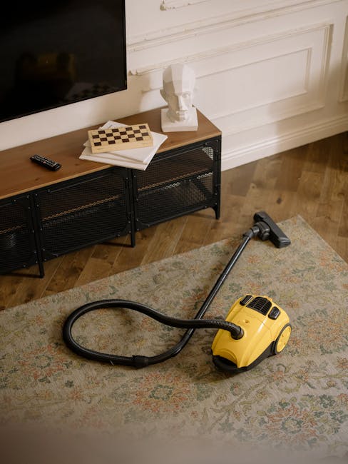 The image depicts a domestic living room scene with a yellow canister vacuum cleaner placed on a patterned rug. The vacuum has a black hose and a cleaning head positioned on the rug, which features a traditional floral design in muted tones. In the background, there is a black metal and wood TV stand holding a flat-screen television, a remote control, a chessboard, and some white papers, all arranged neatly. Above the stand, a decorative white bust sculpture is placed on a wooden side table against a wall with white panel moulding. The wooden floor has a natural finish, and the scene is well-lit, emphasizing the cleanliness and tidiness of the space, illustrating routine surface cleaning and maintenance in a residential setting, as offered by Carpet Cleaning Kingston.