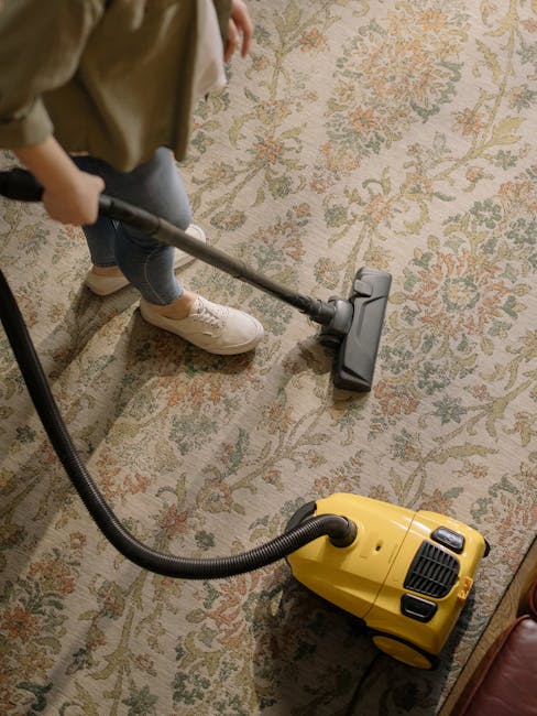 A child using a handheld vacuum cleaner to deep clean a traditional patterned area rug in a residential living room, with a yellow vacuum unit connected by a flexible hose. The rug features floral motifs in muted colors, and the cleaning process appears thorough, with visible suction on the carpet fibers. The room has soft, natural lighting, and the surrounding flooring and furniture are not visible. This scene illustrates domestic surface cleaning, highlighting the importance of maintaining hygiene and freshness in home environments, consistent with services provided by Carpet Cleaning Kingston, as referenced on the page about rug cleaning in Surbiton and Hampton Wick homes.