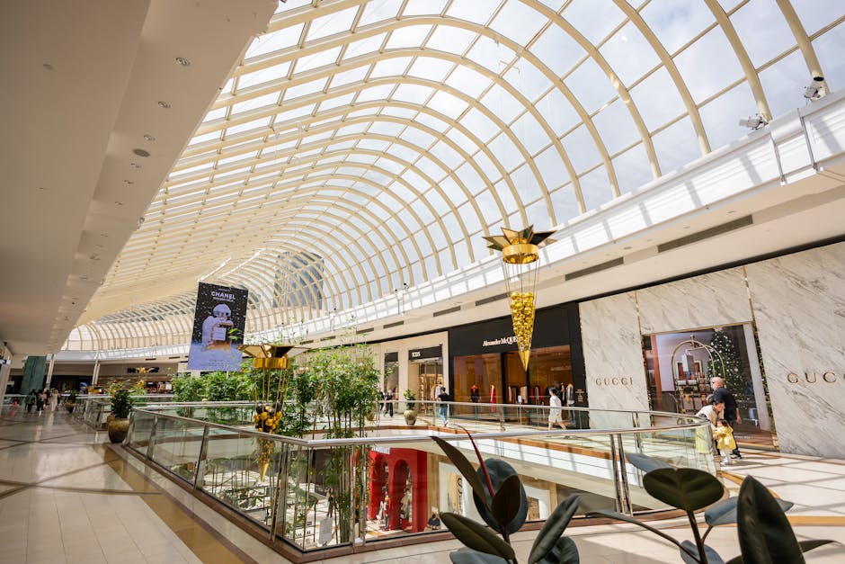 Inside a spacious shopping mall with a high, curved glass ceiling allowing natural daylight to illuminate the interior. The ceiling features a grid of light-colored wooden arches, contrasting with the white ceiling panels. On the upper level, a large digital billboard displays an advertisement for Chanel, with a model holding a bag. Below, the open atrium showcases decorative green plants in large pots, with a glass safety barrier surrounding the central open space. The ground floor has retail storefronts, including Gucci, with elegant white marble walls and wooden accents. The mall floor is made of polished beige tiles, reflecting the bright lighting. A few shoppers are visible walking along the walkway, some carrying shopping bags. Carpet Cleaning Kingston specializes in surface cleaning, deep cleaning, and sanitisation, providing professional cleaning services to maintain the hygiene and cleanliness of retail environments like this one.