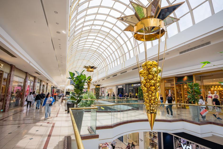 The interior of Bentall Centre shops in Kingston features a spacious, well-lit shopping mall with a high, arched glass ceiling allowing natural light to illuminate the area. The polished tiled flooring appears clean and shiny, reflecting the overhead lighting. Elegant gold and metallic decorations, including a large, cone-shaped hanging ornament filled with gold-colored balls, add a decorative touch to the space. Potted green plants are strategically placed along the walkway, enhancing the fresh and tidy atmosphere. Several retail stores with glass storefronts line both sides of the corridor, displaying various merchandise. Shoppers are casually walking and browsing, indicating a lively but well-maintained commercial environment. The overall appearance suggests routine surface cleaning and maintenance, emphasizing hygiene and aesthetic appeal, a service provided by Carpet Cleaning Kingston as part of their cleaning offerings.