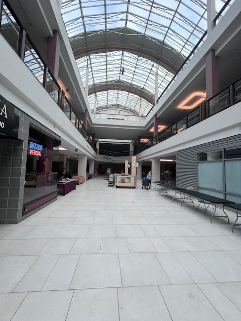 Inside a spacious shopping mall with a high, curved glass ceiling allowing natural daylight to illuminate the interior. The ceiling features a grid of light-colored wooden arches, contrasting with the white ceiling panels. On the upper level, a large digital billboard displays an advertisement for Chanel, with a model holding a bag. Below, the open atrium showcases decorative green plants in large pots, with a glass safety barrier surrounding the central open space. The ground floor has retail storefronts, including Gucci, with elegant white marble walls and wooden accents. The mall floor is made of polished beige tiles, reflecting the bright lighting. A few shoppers are visible walking along the walkway, some carrying shopping bags. Carpet Cleaning Kingston specializes in surface cleaning, deep cleaning, and sanitisation, providing professional cleaning services to maintain the hygiene and cleanliness of retail environments like this one.