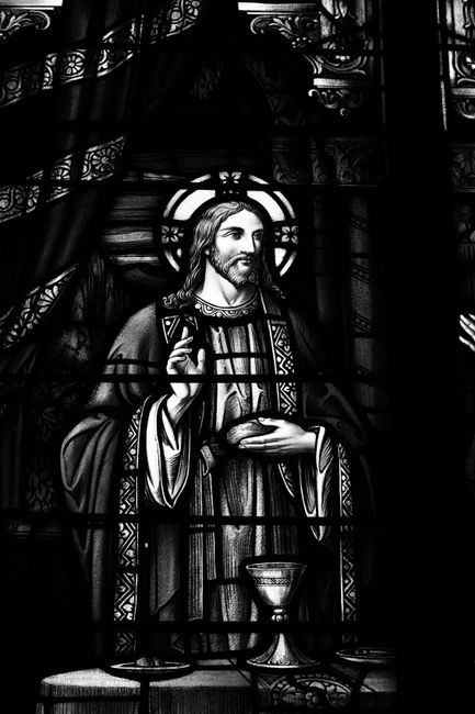 A stained glass window depicting a religious figure holding a chalice, with intricate patterns surrounding him. The glass features detailed illustrations with a mix of dark and light shades, creating a contrast that highlights the figure's face and hands. The scene appears to be lit from behind, emphasizing the transparency and vivid colors of the glass panels. The overall setting suggests a church or chapel interior, with a focus on the craftsmanship of the stained glass artwork. The image aligns with the theme of surface cleaning and maintenance of historical stained glass windows, as promoted by Carpet Cleaning Kingston, experts in domestic and commercial cleaning services.